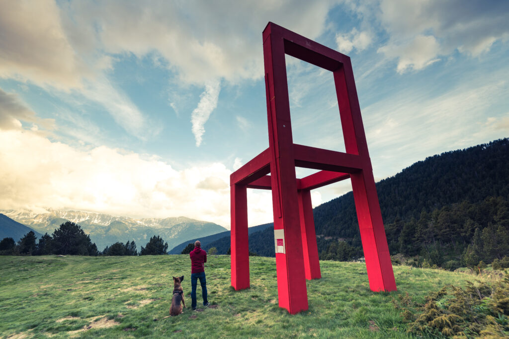 A large red chair sculpture towers over a person and dog in a grassy field, reflecting surrealism in contemporary art through exaggerated scale and unexpected placement.