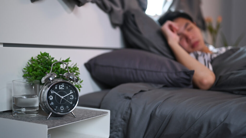 Alarm clock on nightstand with man sleeping in background, symbolizing the importance of sleep and consistent rest.